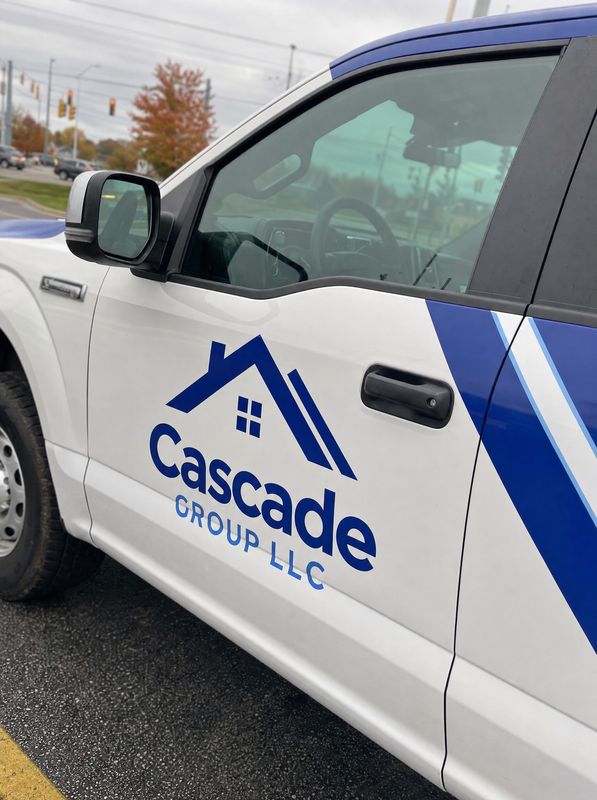 Concept mockup close-up: driver door of a white pickup with a navy house-roof icon, navy "Cascade" and blue "Group LLC" lettering, and the edge of the blue stripe graphics.