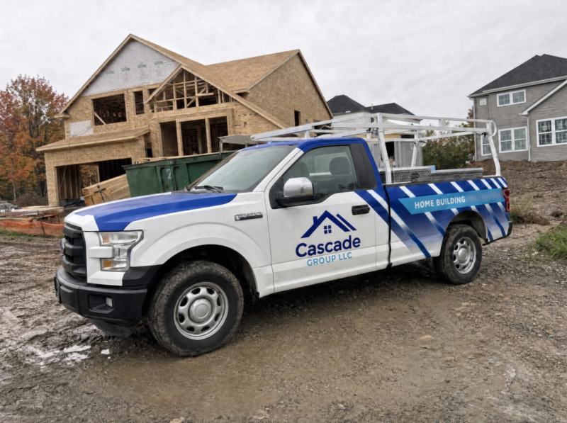 Concept mockup: white Ford F-150 pickup, front three-quarter view, parked on a muddy new-home construction site, showing the blue hood stripe, the "Cascade Group LLC" door logo, and the blue striped bed wrap with a "Home Building" banner.